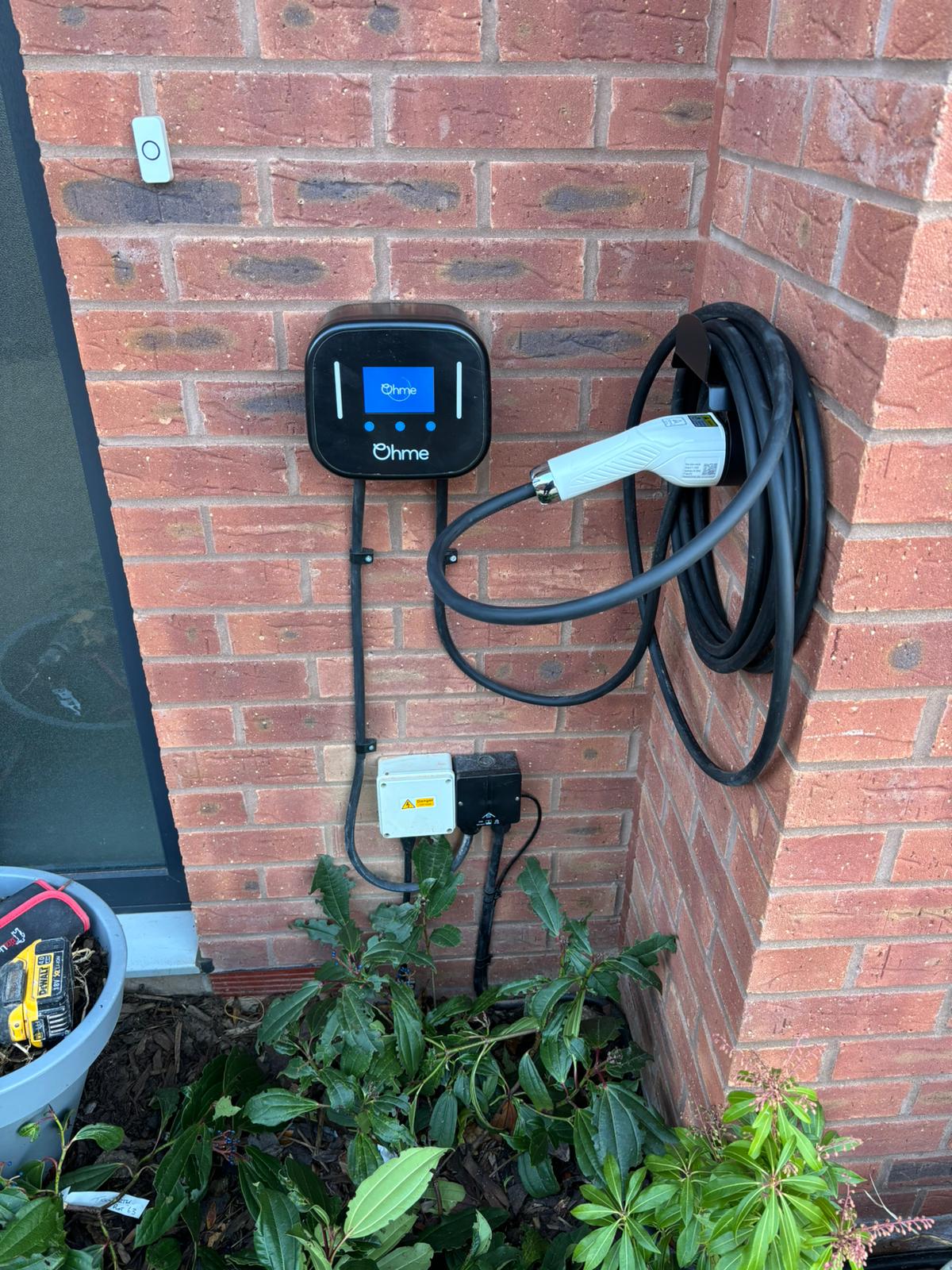 A professionally installed Ohme EV charger mounted on a red brick wall outside a home. The charger is securely connected with electrical wiring running down to a protective junction box. The charging cable is neatly coiled on a wall-mounted holder. Green plants grow near the base of the installation, and a toolbox with equipment is visible in the lower left corner.